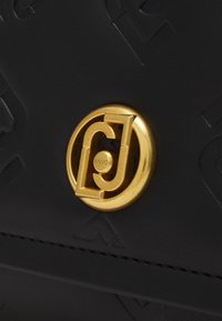 Borsa a tracolla in pelle nera con texture, caratterizzata da una chiusura circolare dorata con un dettaglio a logo a strati e un motivo in rilievo sulla superficie.