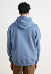 Felpa blu realizzata in morbido cotone misto, con un cappuccio ampio, polsini e orlo a costine. Indossata sopra pantaloni in colori neutri.