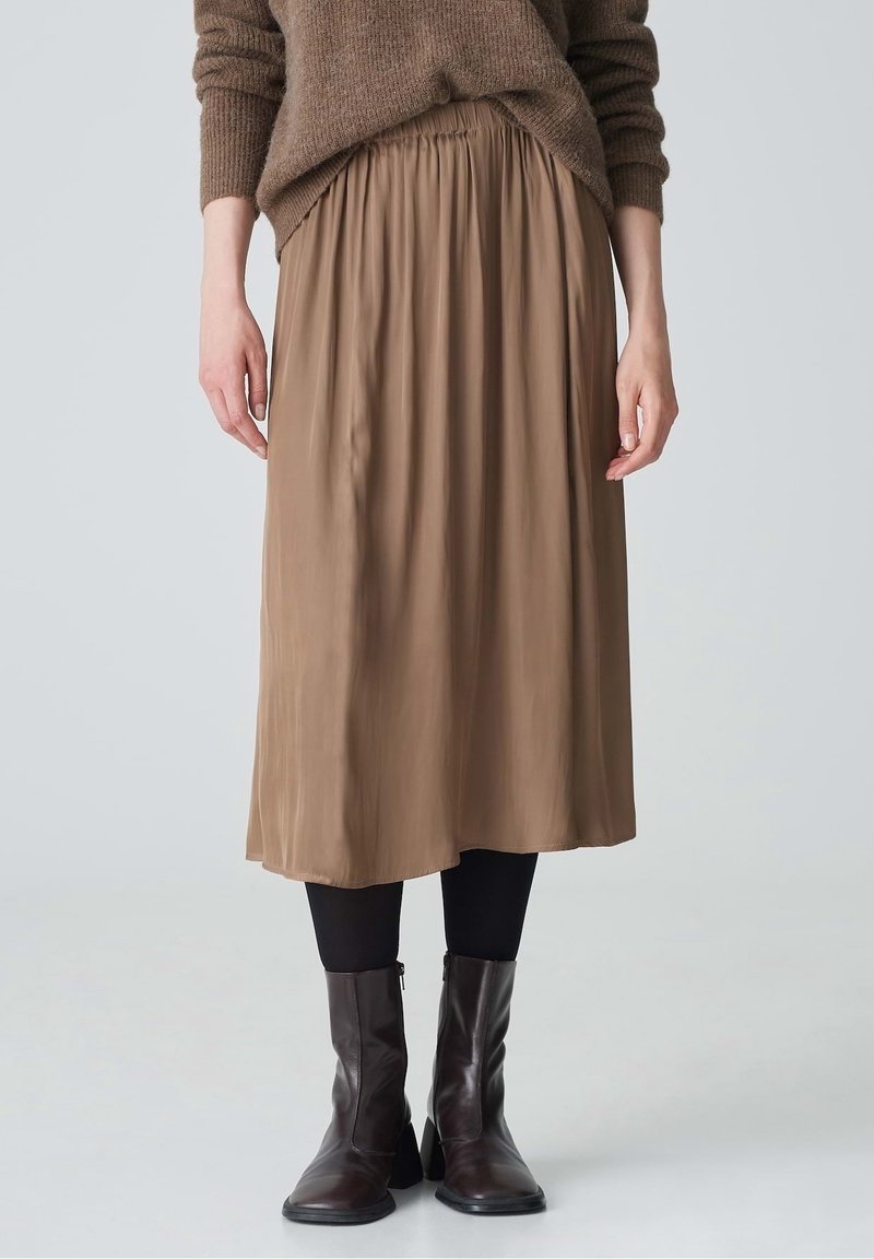 Personne portant une jupe marron jusqu'aux genoux, un pull marron, des leggings noirs et des bottines marron foncé sur un fond clair uni.