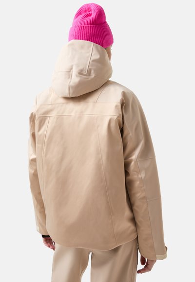 Beige waterdichte jas met capuchon, met een gladde textuur en gestikte panddetails. Gecombineerd met een felroze gebreide muts.