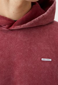 Gros plan d'une personne portant un sweat à capuche rouge Under Armour montrant la capuche pliée et l'écusson du logo sur la poitrine.