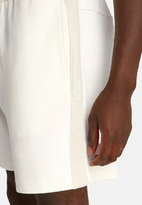 Bermudas de algodón blancas con una franja lateral beige texturizada, con el logo de "Calvin Klein" en una fuente sutil en la cinturilla.