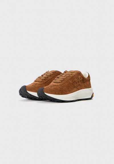 Adidași din suede maro cu sireturi bej, tălpi intermediare albe și tălpi exterioare texturate negre, având un logo cusut pe lateral.