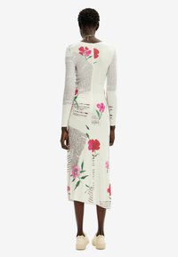 Robe en tricot à manches longues d'un crème clair, avec des motifs floraux et des impressions textuelles. Longueur midi avec une coupe droite et un détail fendu subtil.