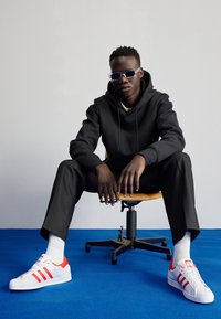 Zapatillas Adidas blancas con rayas rojas, sudadera negra, pantalones negros, sentado en una silla contra un fondo liso y una alfombra azul.