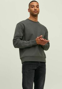 Dark green sweatshirt, crew neck, soft fabric, ribbed cuffs and hem, paired with dark jeans. Model stands with hands together, neutral background.