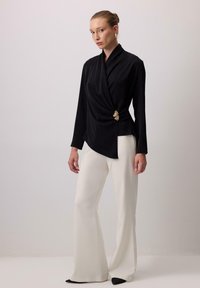 Melnā wrap blūze, kas izgatavota no mīksta auduma ar garām piedurknēm, ar zelta akcentu. Apvienota ar baltām platām biksēm, piedāvājot elegantu siluetu.