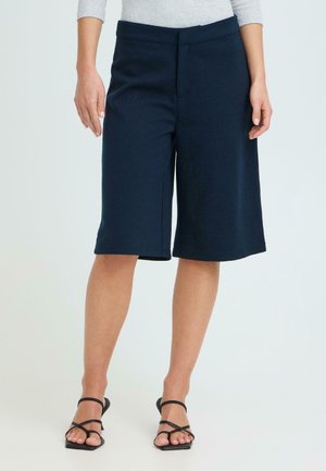 Vrouw die marineblauwe wijde shorts tot op de knie draagt met zwarte sandalen met hakken en bandjes, staand tegen een effen lichte achtergrond.