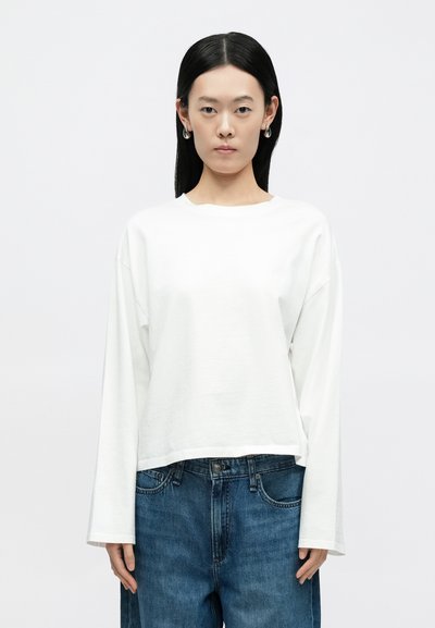 Biała koszula z długim rękawem wykonana z miękkiej bawełny, o luźnym kroju i szerokim dekolcie, zestawiona z niebieskimi dżinsami. Model stoi zwrócony przodem.