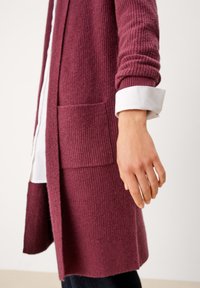 Cardigan lungo color bordeaux realizzato in tessuto a coste, con due tasche frontali e polsini di camicia bianchi arrotolati. Texture morbida e orlo dritto.