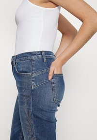 Jeans de mezclilla azules con un corte ajustado, un bolsillo lateral y detalles decorativos a lo largo de la pierna. La tela tiene una textura y acabado clásico.