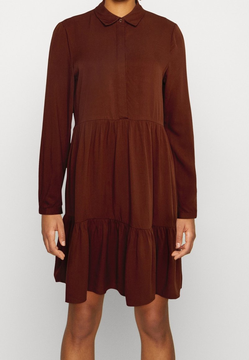 Robe marron à manches longues avec col et jupe à volants, portée par une personne debout devant un fond uni, les mains détendues sur les côtés.