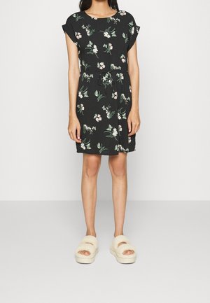 Robe courte noire à motifs floraux verts et roses, col rond et taille à nouer. Portée avec des sandales à glissière crémeuses et duveteuses.