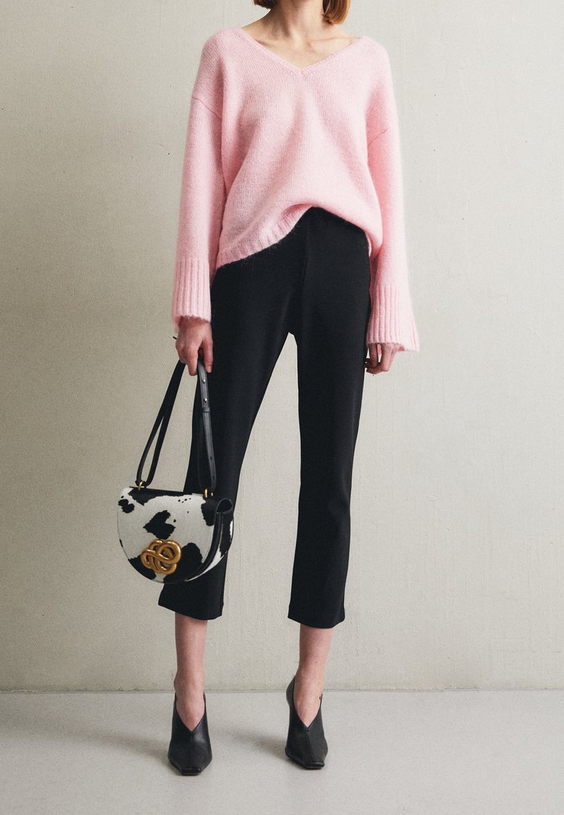 Femme portant un pull rose ample à col en V, un pantalon court noir, des chaussures noires pointues, tenant un sac à main noir et blanc à motif.