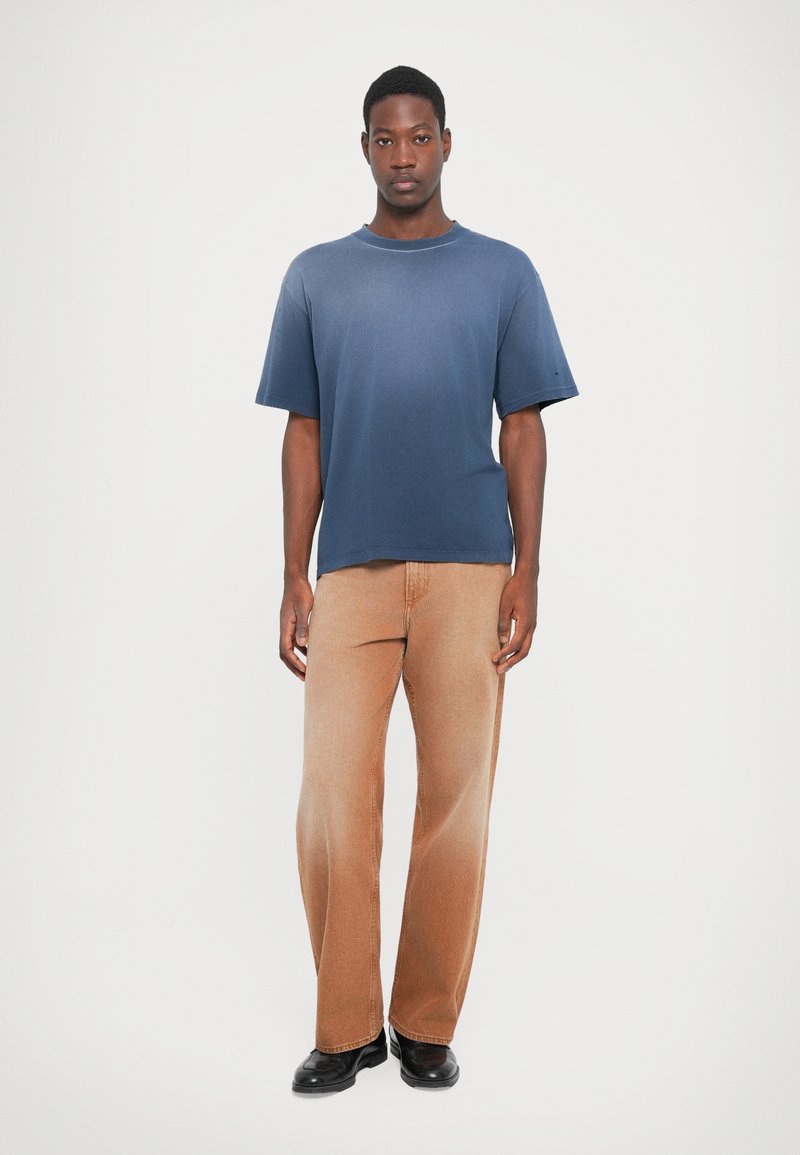 Marineblaue kurzärmlige T-Shirt mit Rundhalsausschnitt, kombiniert mit hellbraunen, geraden Hosen und schwarzen Schuhen auf einem einfarbigen Hintergrund.