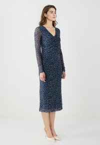 Abito da sera blu navy con una forma aderente, maniche lunghe in tessuto trasparente, scollatura a V e dettaglio frontale arricciato. Presenta un motivo a pois multicolore su tutta la superficie.