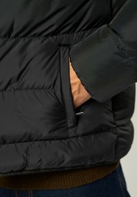 Svart dunjakke med quiltet tekstur, med en sidelomme. Materialet ser glatt og isolert ut, egnet for kaldt vær.