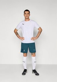 Vyrų sportininkas stovi su rankomis ant klubų, vilkėdamas baltą Puma marškinėlį, mėlynas šortus su numeriu 1, baltas iki kelių čėsnus ir juodus batus.