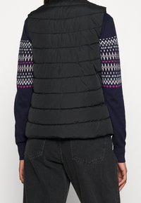 Person som bär en svart quiltad ärmlös väst över en marinblå tröja med vita och rosa geometriska mönster, samt mörkgrå jeans.