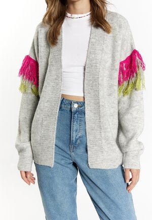 Femme portant un cardigan gris clair avec des franges roses et vertes aux manches, un top blanc court et un jean bleu taille haute.