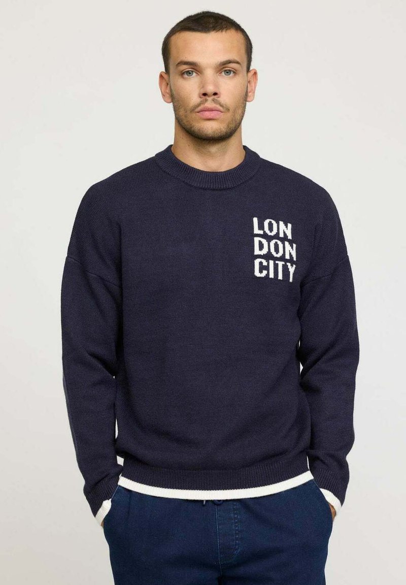 Pull en maille marine avec un col rond et du texte blanc "LONDON CITY" sur la poitrine. Présente des poignets et un ourlet côtelés avec une bordure blanche contrastante.