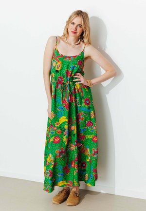 Femme blonde portant une robe maxi sans manches à motifs floraux et de fruits verts, chaussures marron sans lacets, et colliers superposés, debout contre un mur blanc.
