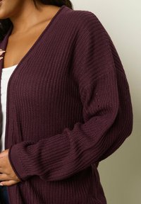 Bordeaux neulecardigan, jossa on löysä istuvuus, syvä V-pääntie, ribbikuviointi ja pitkät hihat. Hihassa on hienostuneita yksityiskohtia.