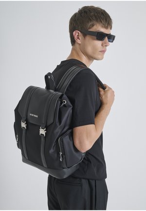 Mochila negra hecha de material sintético suave, con un diseño estructurado, dos asas superiores, herrajes plateados y bolsillos con cremallera en el exterior.