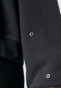 Tessuto di felpa nera con una texture liscia, caratterizzato da un bordo grezzo e un dettaglio con bottoni a pressione metallici sulla manica.