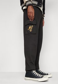 Schwarze Jogginghose mit einer seitlichen Cargo-Tasche, die mit goldfarbenem "NE" bestickt ist. Der Stoff hat eine glatte Textur. Kombiniert mit schwarzen High-Top-Sneakers.