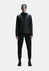 Black lightweight jacket with a hood, zip closure, and side pockets, paired with matching black tapered pants and black athletic shoes.