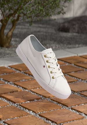 Biały niski sneaker z złotymi oczkami i oznaczeniem na drewnianych kafelkach na zewnątrz, w tle widoczne drzewo i kamienna podłoga.