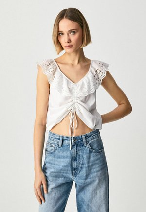 Top de algodón blanco con escote en V y mangas de encaje con volantes. Presenta un detalle de cordón en la parte frontal y un corte corto. Combinado con jeans azules.