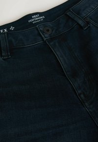 Tumesinised teksapüksid sirge lõikega, ees nööpidega ja toonitoonilise õmblusega. Sildil on kirjas "NEXT PERFORMANCE."