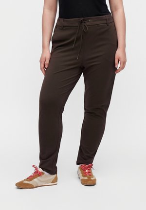 Person iført mørkebrune snøre bukser med beige og brune sneakers med lyserøde snørebånd, står mod en ensfarvet hvid baggrund.