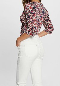 Blouse à imprimé floral avec manches bouffantes, présentant des nuances de bleu marine, rouge et crème ; associée à un jean blanc taille haute avec des accents dorés.
