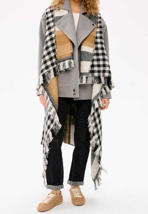 Henkilö, joka pukeutunut harmaaseen takkiin, mustiin käännettyihin farkkuihin, vaaleanruskeisiin tennareihin ja suureen mustavalko-keltaiseen ruudulliseen, hapsulliseen huiviin.