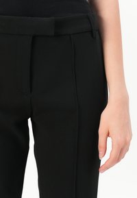 Zwarte tapershorts met een gladde textuur, voorzien van een tailleband, voorkeursplooien en zijzakken. Close-up van een hand die de zijzak vasthoudt.