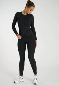 Czarny top z długim rękawem w połączeniu z dopasowanymi czarnymi legginsami, wykonany z elastycznego materiału. Uzupełniony białymi i zielonymi butami sportowymi. Prosty design.