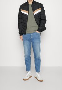 Černá prošívaná bomber bunda s hnědými, bílými a černými šipkovými pruhy; olivově zelená košile; světle modré džíny; bílé tenisky s hnědými podrážkami.