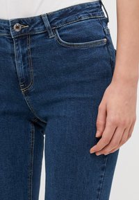 Jeans de mezclilla azul oscuro con un diseño ajustado, costuras amarillas contrastantes y cierre frontal con botón. Los bolsillos son visibles a los lados.