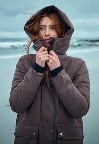 Brauner wasserdichter Parka mit Kapuze, ausgestattet mit einem Reißverschluss und Druckknopfverschluss, seitlichen Taschen und einer glatten Textur, getragen von einer Person im Freien.