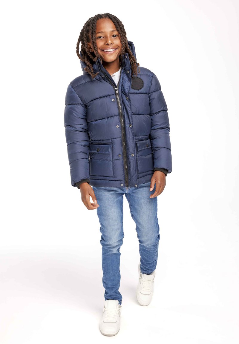 Pufjakas tumši zilā krāsā, ar rāvējslēdzēju priekšpusē, divām priekšējām kabatām un kapuci, apvienots ar gaiši zilām džinsām un baltām sporta apaviem.