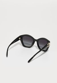 Lunettes de soleil noires avec une monture épaisse et brillante, verres gris dégradés et accents dorés sur les tempes, présentant une forme moderne de chat.