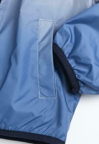 Veste légère bleue avec un extérieur lisse, des poignets élastiques, un bord contrastant marine et une poche verticale sur le côté gauche.