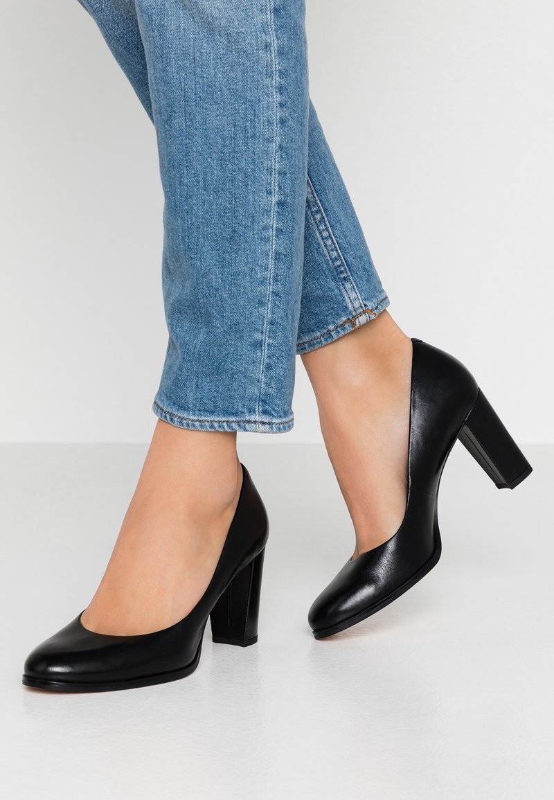 Escarpins à talons hauts en cuir noir avec un bout arrondi, un talon bloc et une texture lisse, portés avec un jean denim bleu raccourci.