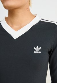 Camisola preta de mangas longas com colarinho branco e o logótipo da Adidas. Apresenta um design em V e riscas brancas horizontais nas mangas.