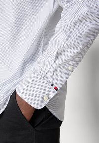 Camicia bianca con un piccolo motivo a croce blu navy, caratterizzata da un polsino con bottoni e un accento di cucitura in rosso, bianco e blu.