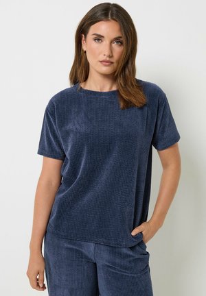 Vrouw met stijl bruin haar die een losse donkerblauwe getextureerde top met korte mouwen en bijpassende broek draagt, één hand in de zak, neutrale uitdrukking.