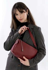 Burgunderrotes Lederhandtasche mit einer strukturierten Form, verstellbarem Griff und dekorativen Hardware-Details. Glatte Textur und minimalistisches Design.